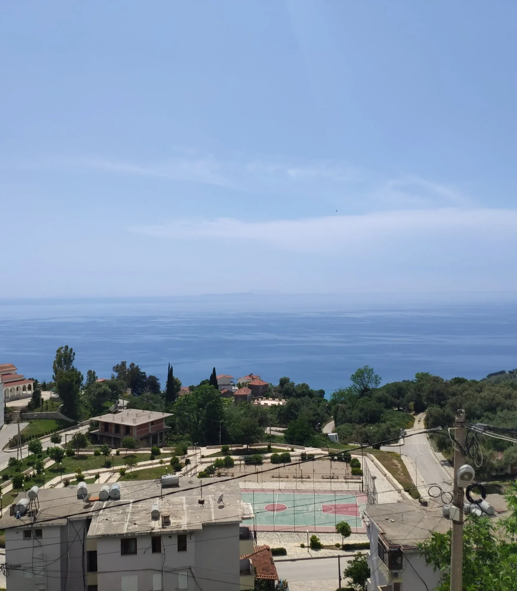 Elevated daytime view of the Lukovë village church, local sports court, and the clear blue sea horizon.
