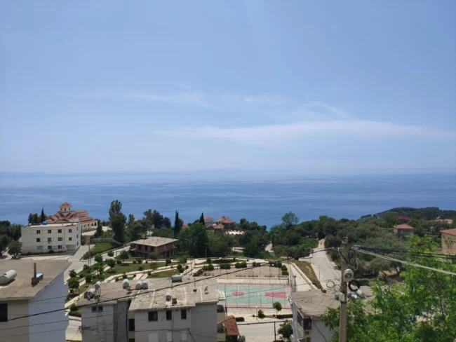 Elevated daytime view of the Lukovë village church, local sports court, and the clear blue sea horizon.