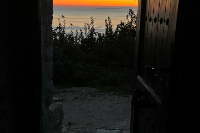 Golden sunset and ocean horizon framed by the open wooden door of the historic Shën e Premte stone church.
