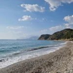Gentle ocean waves washing onto the pebble shore of Lumra Beach with coastal mountains in the background in Lukovë.